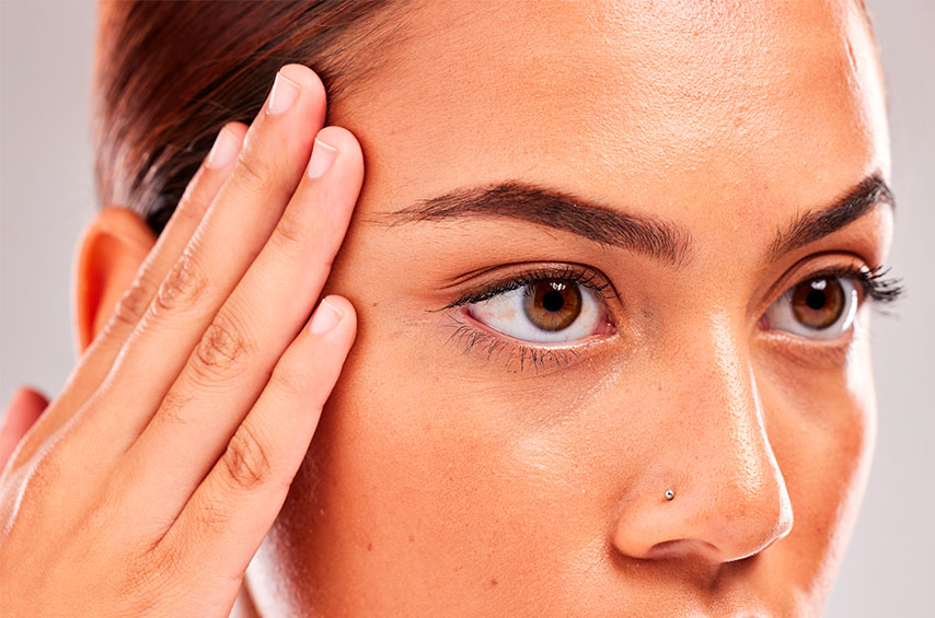 A imagem é um close nos olhos de uma mulher de pele bronzeada e olhos castanhos que olha para além da câmera. Sua mão direita está posicionada ao lado de sua sobrancelha de mesmo lado.